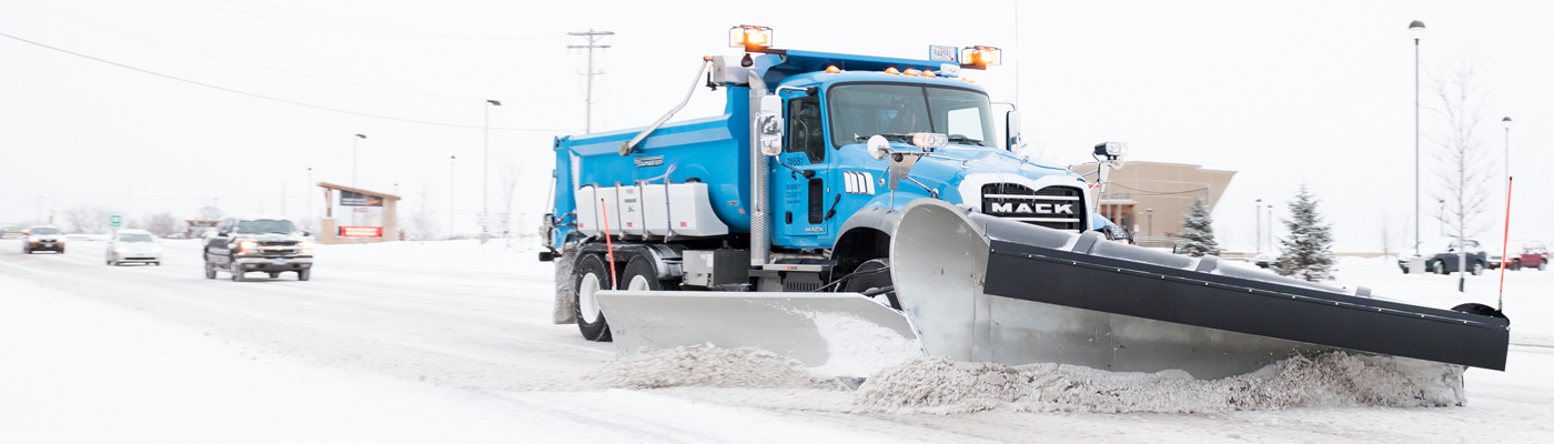 Blue snow plow plows a road with several vehicles following from a distance.