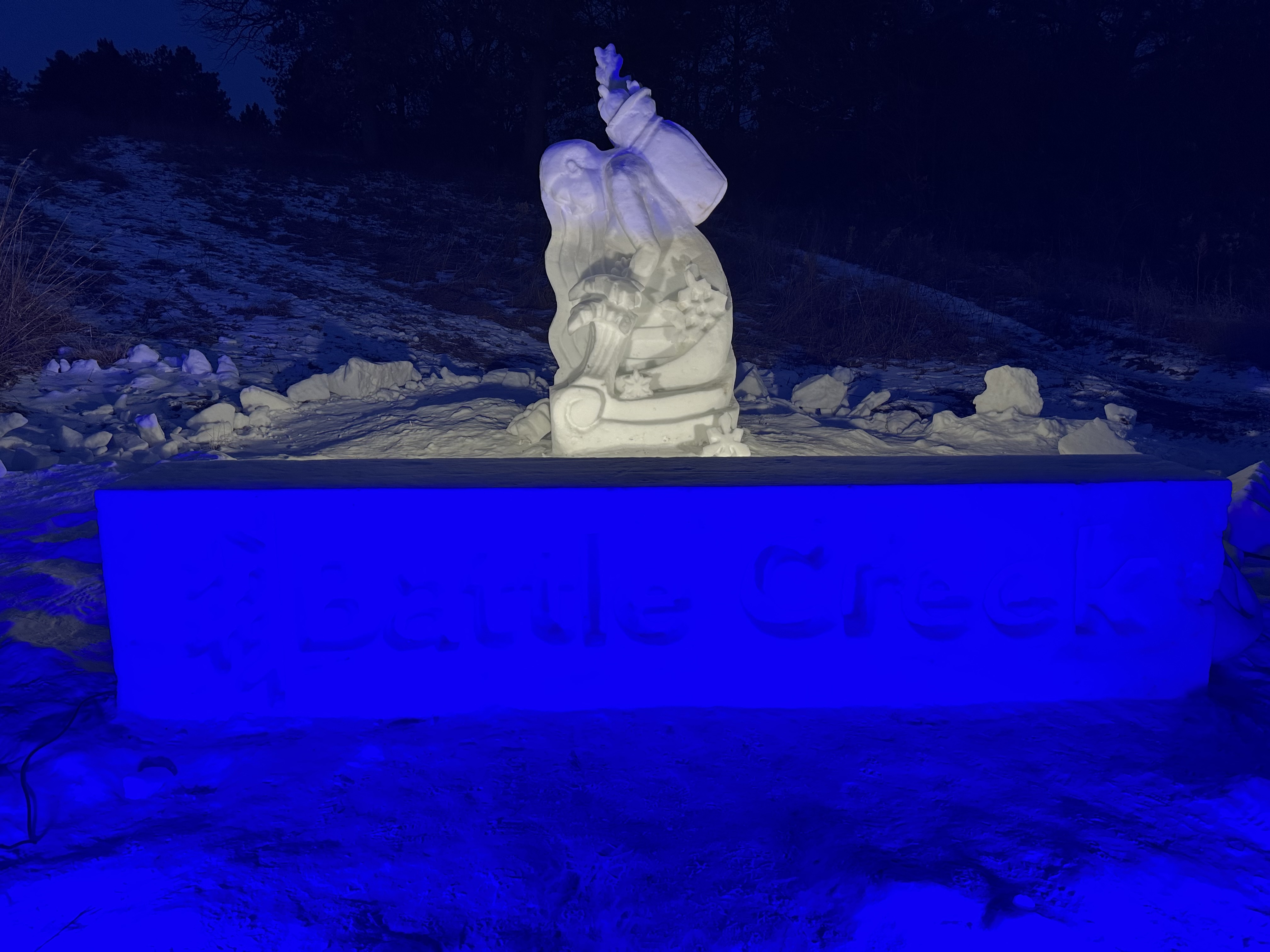 Snow sculpture of a woman kneeling in the snow at the Ramsey county Battle Creek Winter Recreation. Sculpture featured at night with a blue light.