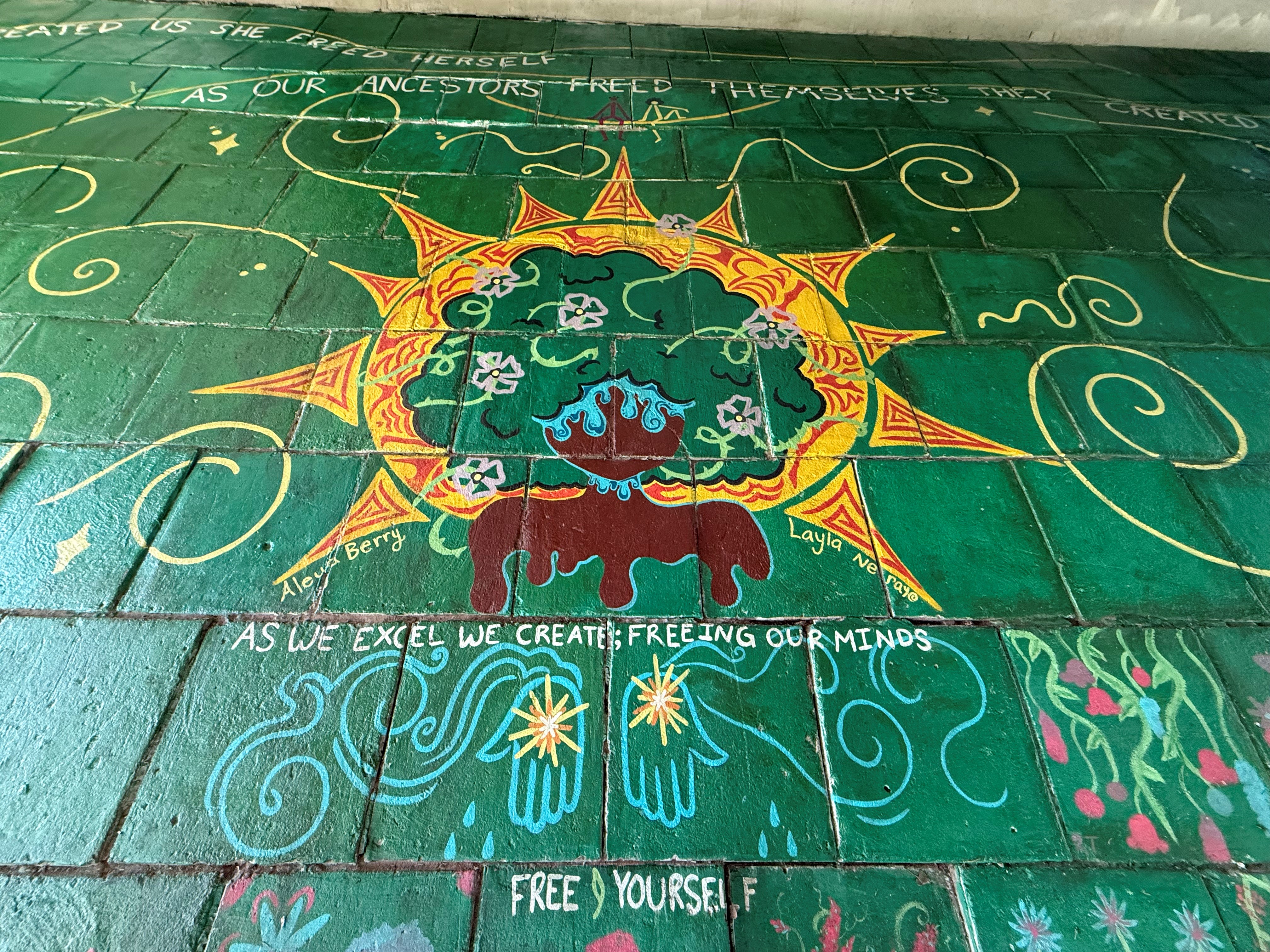 Mural painted on green bricks. Art depicts a black woman encased in a yellow and red sun. She is surrounded by brightly colored flowers and words. Words read "as we excel we create; freeing our minds." artist Aleya Berry Layla Nerayo 