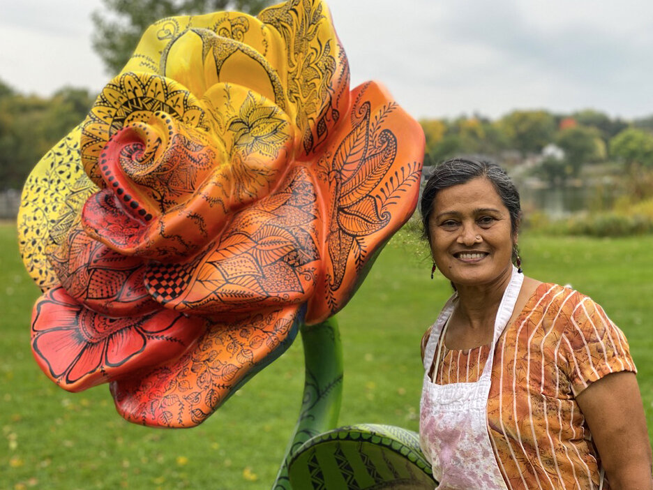 Artist Shakun Maheshwari smiling next to her yellow, orange, and red colored rose sculpture at the park.