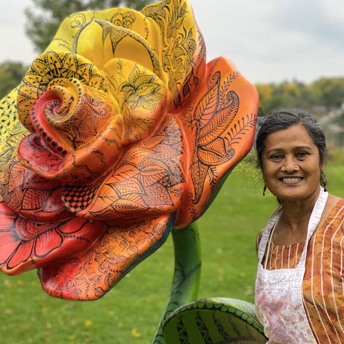 Artist Shakun Maheshwari smiling next to her yellow, orange, and red colored rose sculpture at the park.