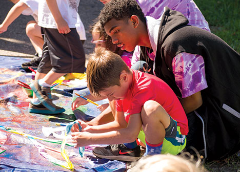 A Junior Assistant Naturalist volunteer assisting a day camp participant with an outdoor craft activity.