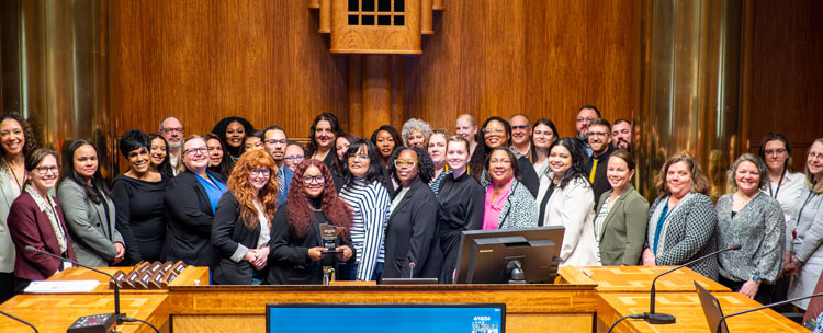 36 people posing in the Chambers. A woman near the middle is holding a Minnesota-shaped plaque.