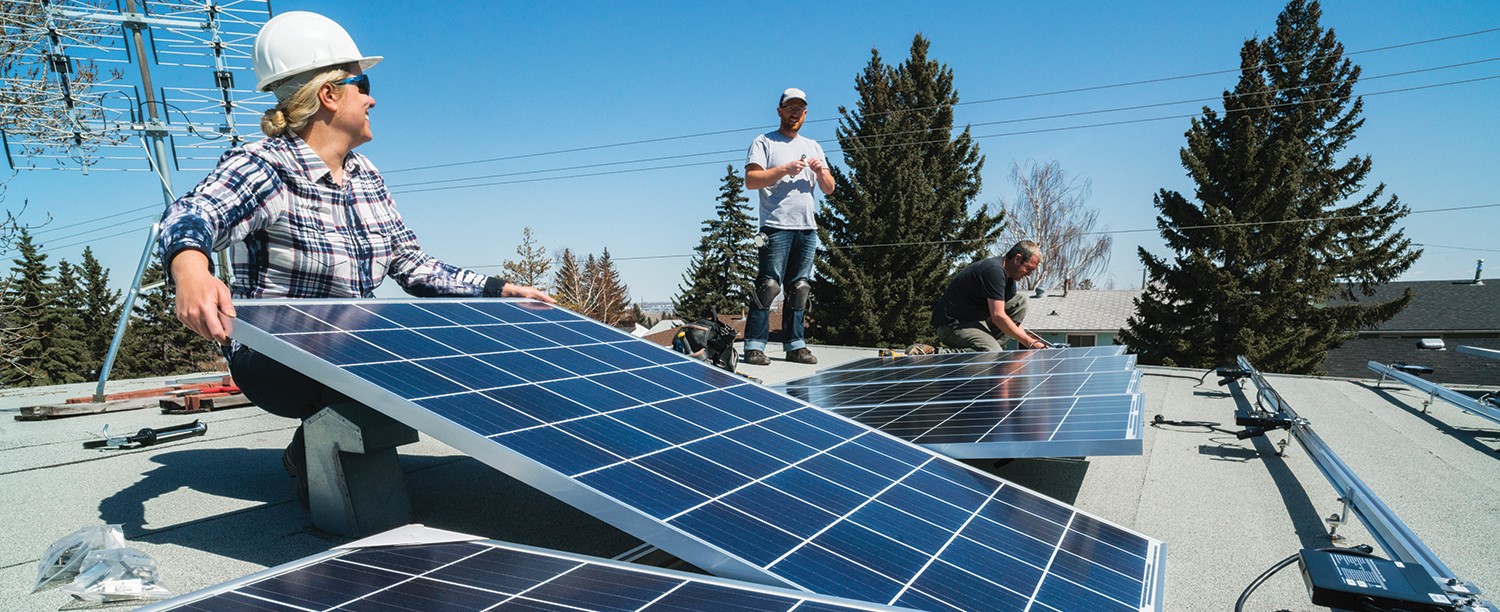 A person wearing a hard hat holds up a solar panel on the roof of a building. Another worker in a hard hat is visible in the background. A bright blue sky is also visible.