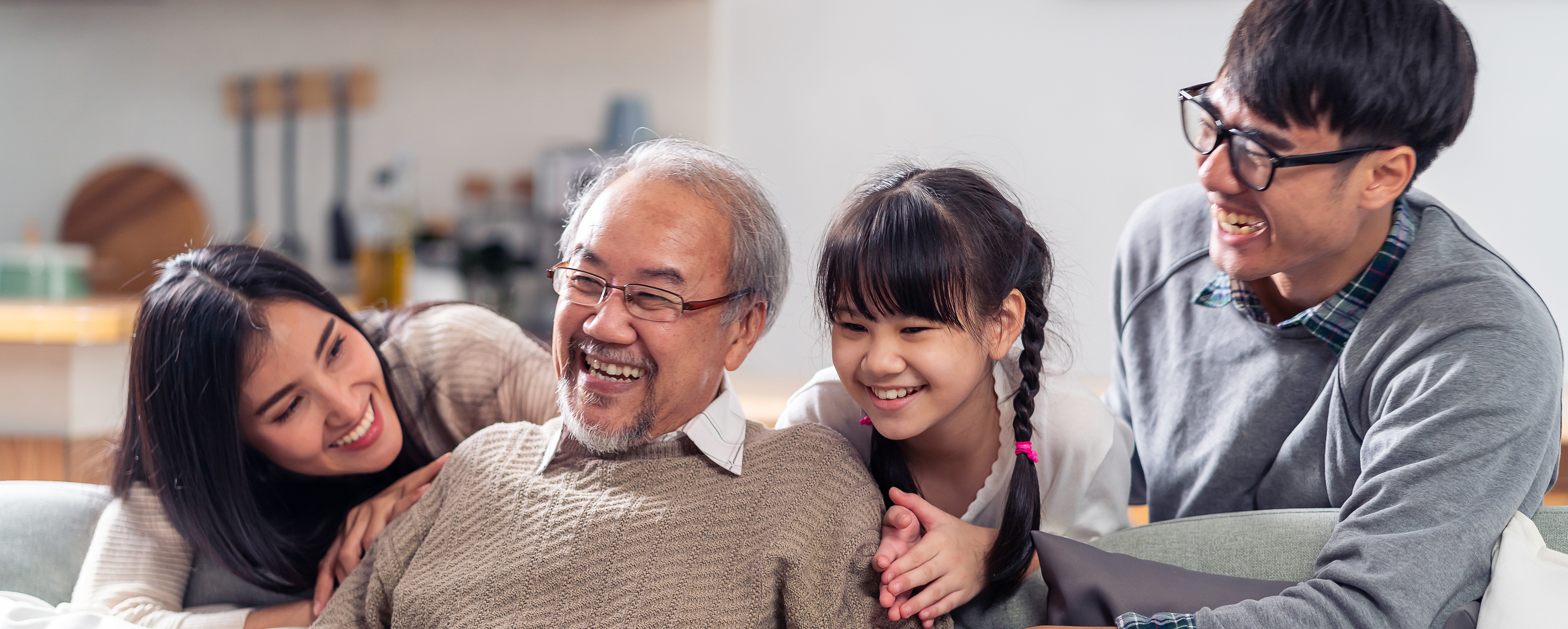 An elderly man sits on a white couch. An adult woman, a girl, and an adult man wearing glasses lean over the back of the couch to embrace him.