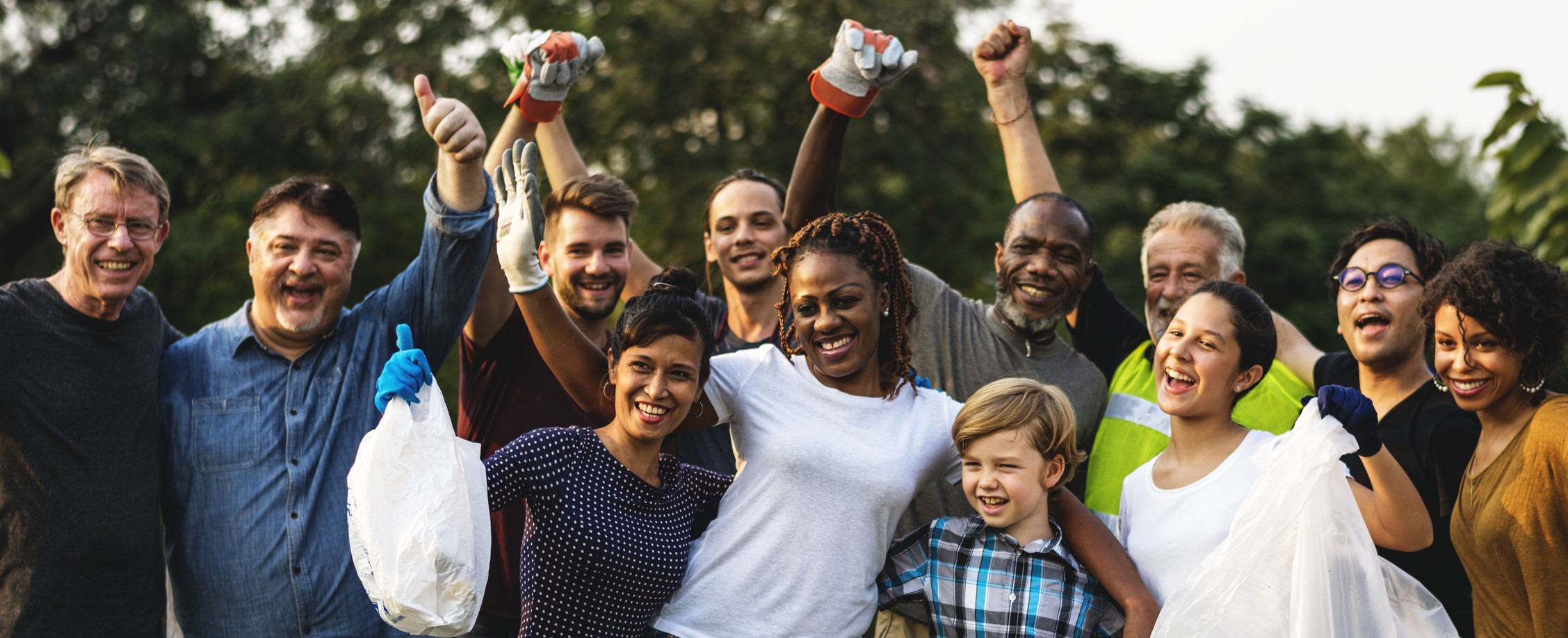 Image: A group of people stand together outside. Some have their arms in the air triumphantly. All are smiling. Some wear gardening gloves. Some are holding white plastic bags.