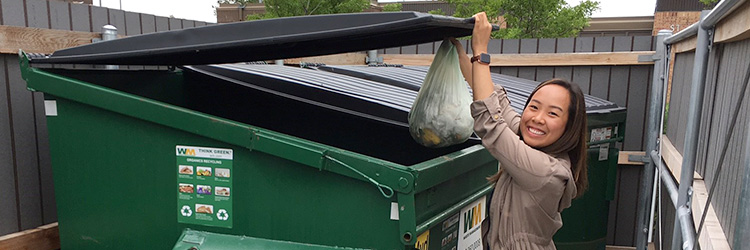 Woman placing a bag of food scraps into a green food scraps drop-off site dumpster.