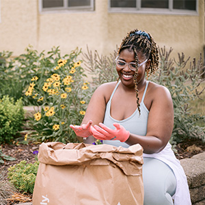 Woman wearing gardening gloves smiles while sitting behind a large paper bag of gardening waste.