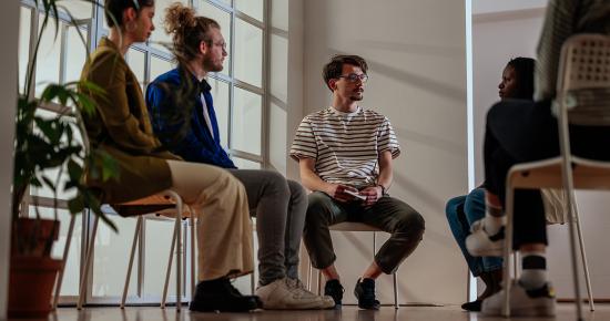 A group of adults sitting in chairs organized in a circle. Everyone is looking towards one person who is speaking