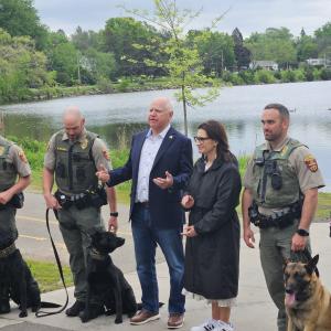 Govenor Walz, Lieutenant Governor Flanagan, DNR Commissioner Sarah Strommen, and four DNR officers along with their DNR sniffing dogs are posed in a photo in front of a lake. 