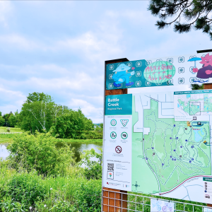 Battle Creek Recreation Trail sign that depicts a map of the area. On top of the map is artwork done by CIRCE of a red and blue caricature on either side with the words in the middle saying "rain or shine for all kind."