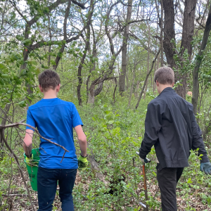 Two boys seen from behind walking in a wooded area with gloves on. One boy has a green bucket in hand and the other boy has a grey shovel.
